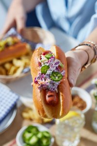 A hand holding out a hot dog with coleslaw and jalapeño toppings.